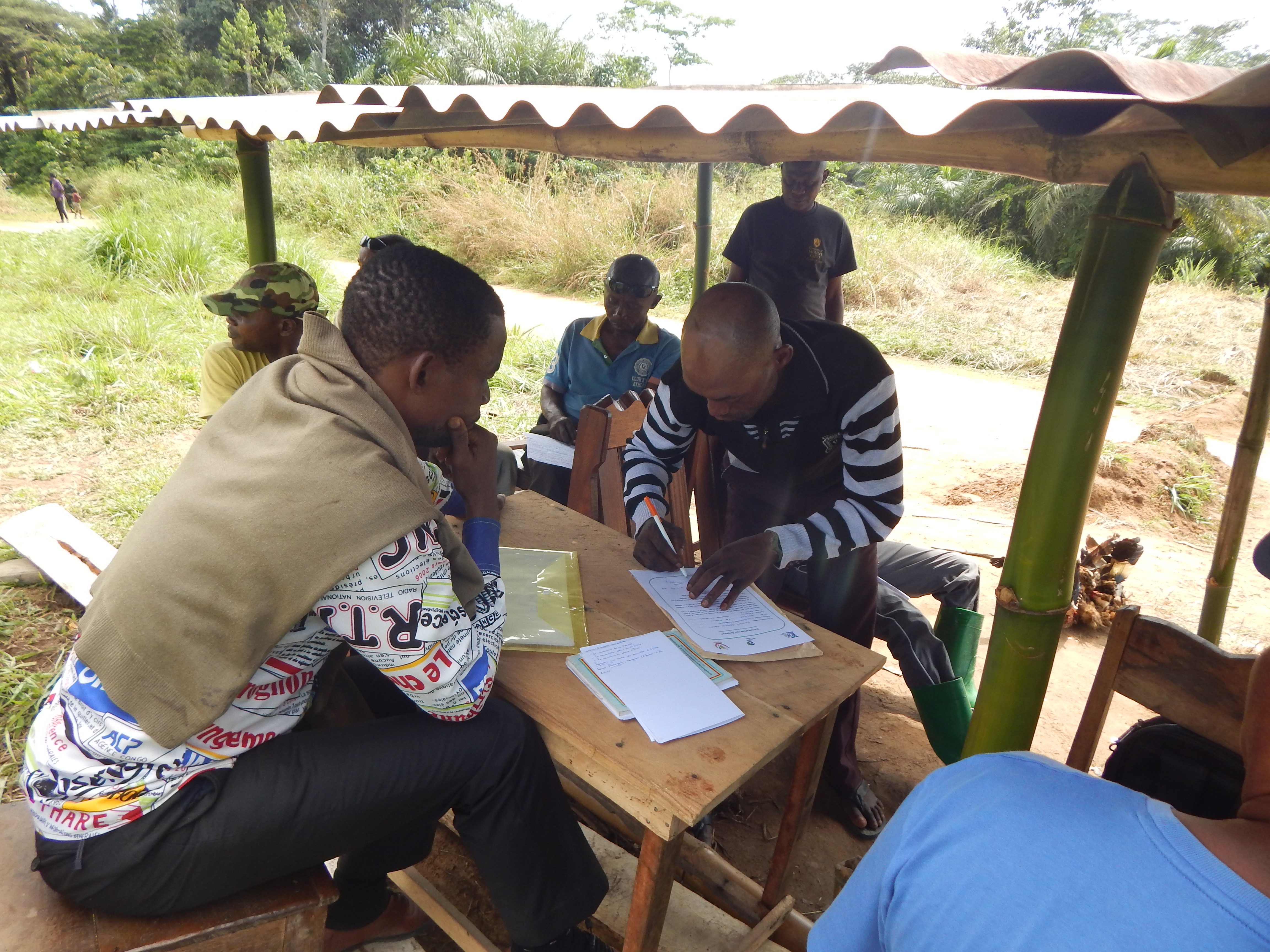 A man signs a document at a table under a tin roof while another man watches.